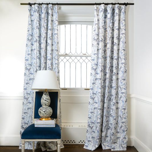 Blue With Intricate Tiger Design Printed Curtains on black rod in front of an illuminated window with Navy Blue Velvet chair with white lamp with designs on top of stacked books