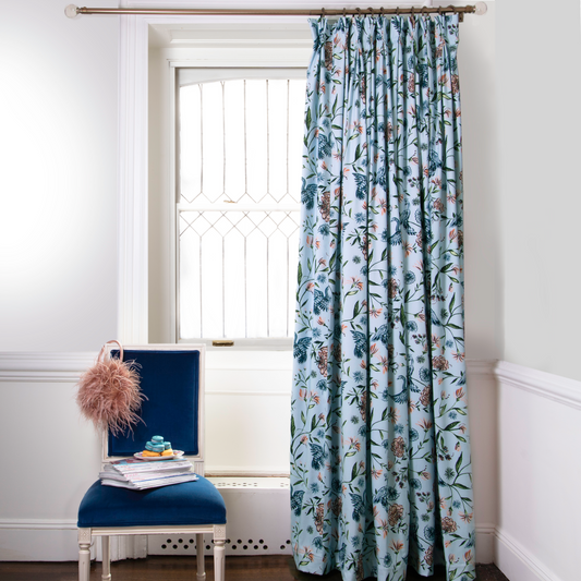 Blue Chinoiserie printed curtains on metal rod in front of an illuminated window with navy chair with coral pom pom hanging in front stacked with books and macaroon plate