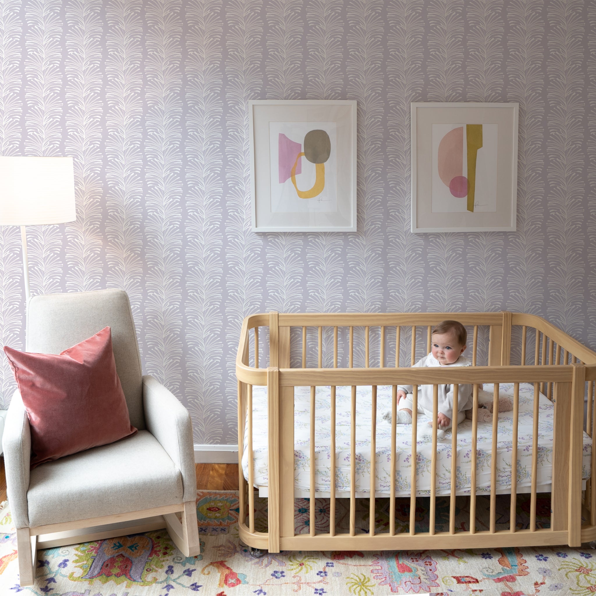 Nursery room with Lavender Botanical Stripe Printed Wallpaper with two art frames on top of crib with baby on top next to White armchair with Coral Velvet Pillow on top next to White tall lamp 
