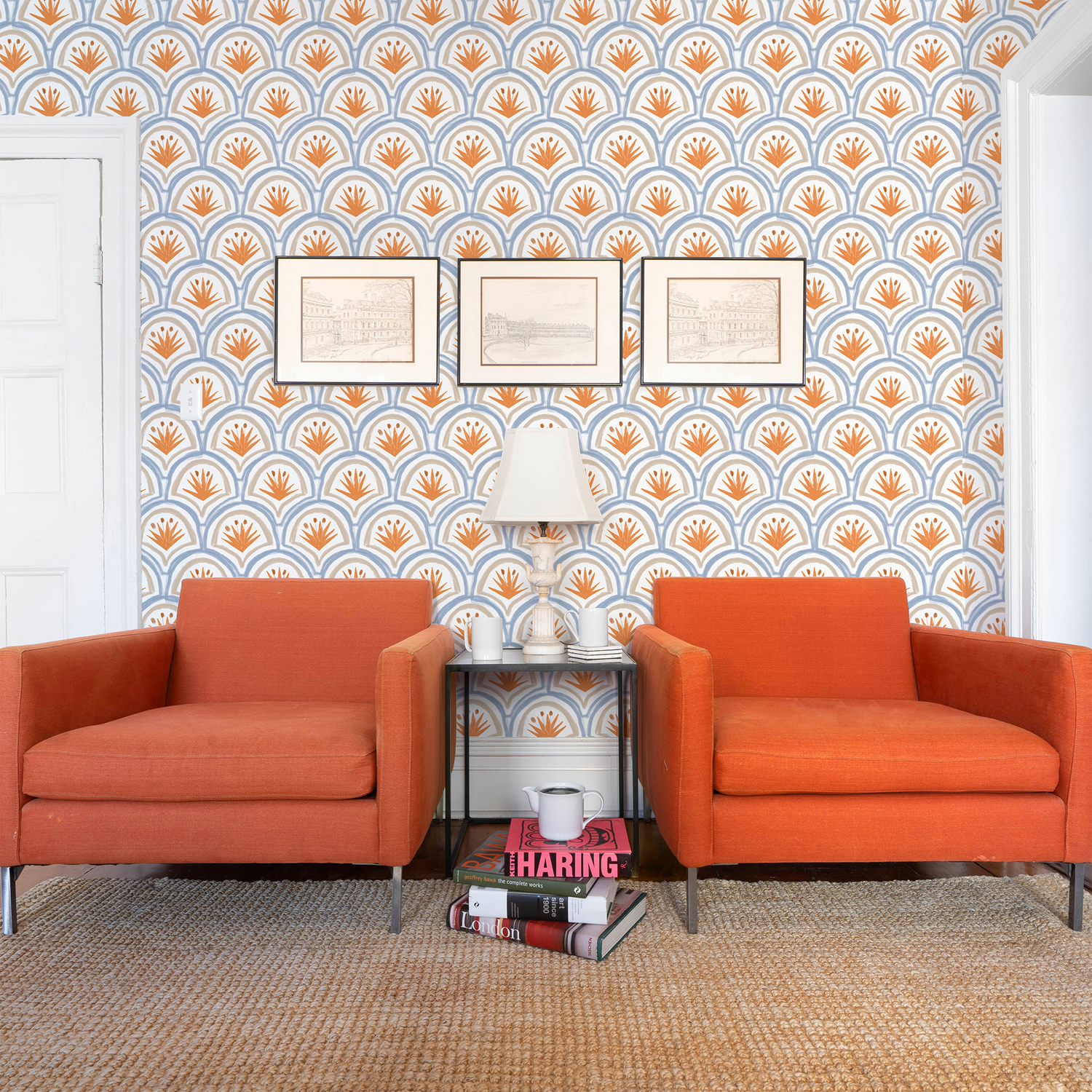 Living room styled with Art Deco Palm Pattern Printed Wallpaper and two orange sofa chairs with a white lamp on table and books stacked on floor in between them