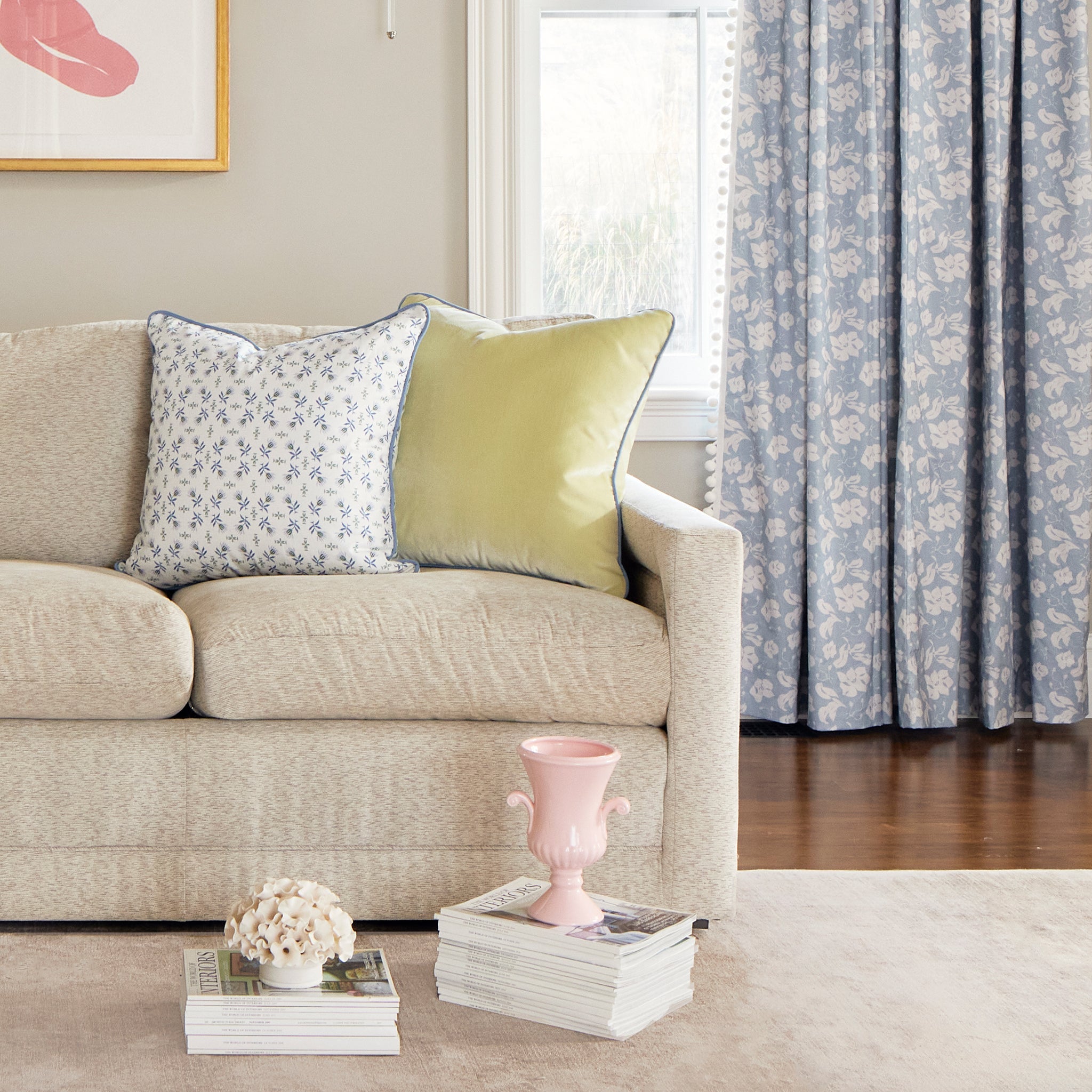 Living room corner styled with Cornflower Blue Floral Printed Curtains with White Pom Poms and cream couch with Blue & Green Floral Printed Pillow and Light Green Pillow behind two stacks of magazines with two pink decorations on top