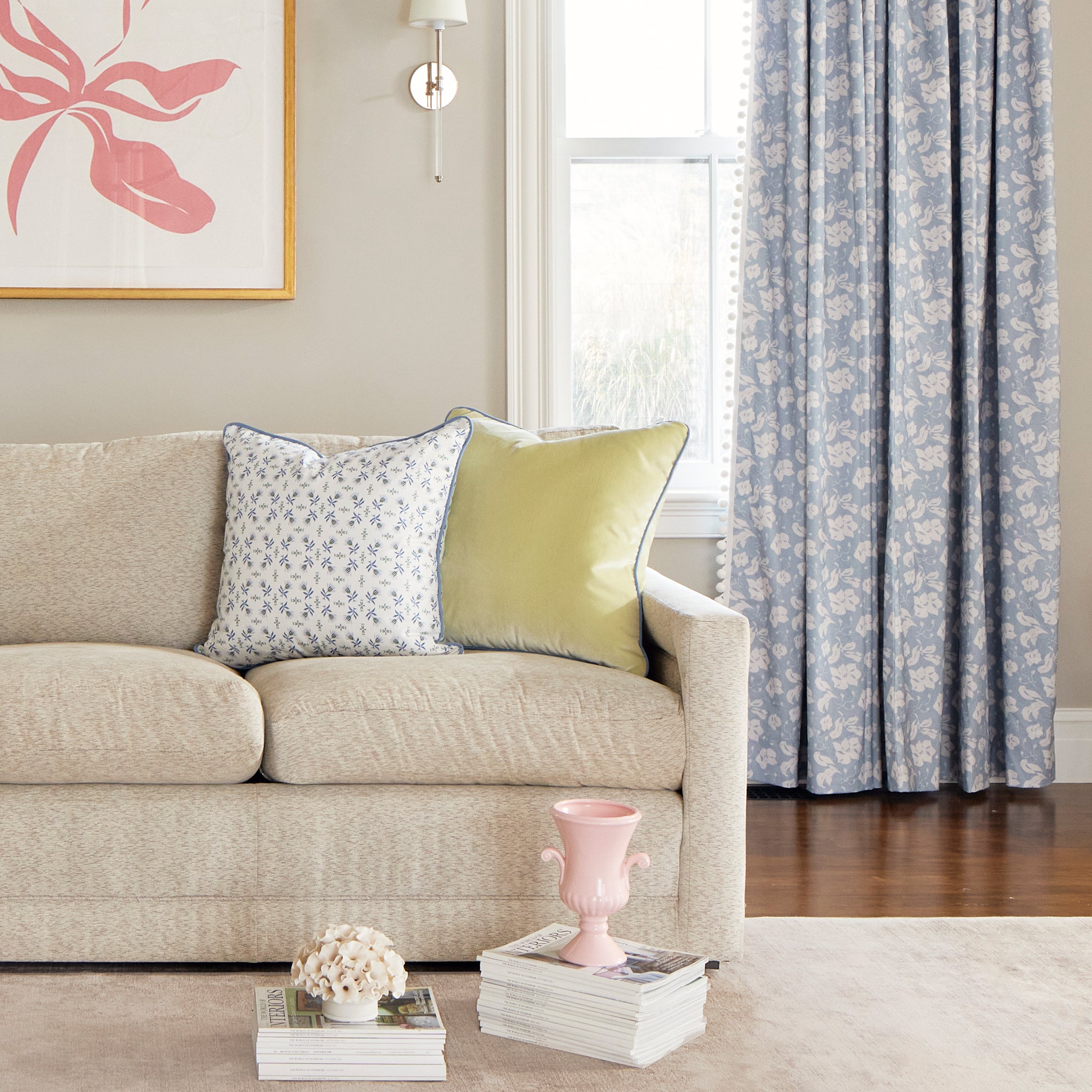 Living room corner styled with Cornflower Blue Floral Printed Curtains with White Pom Poms and cream couch with Blue & Green Floral Printed Pillow and Light Green Pillow behind two stacks of magazines with two pink decorations on top