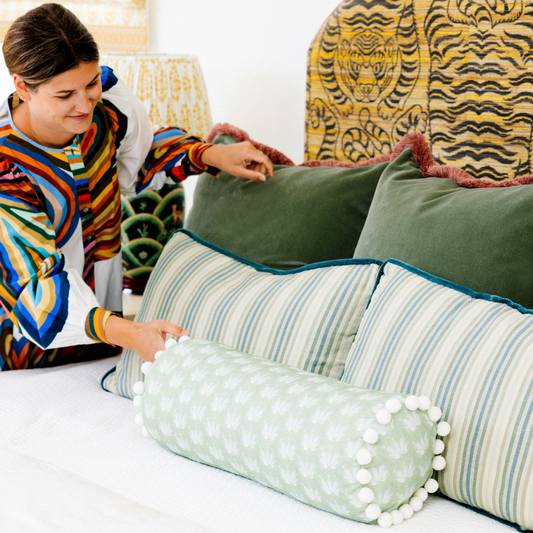 Brunette woman holding one fern green velvet custom pillow next to one other with two striped lumbar pillows in front and a Blue & Green Floral Drop Repeat custom bolster pillow with white pom poms over a tiger printed headboard