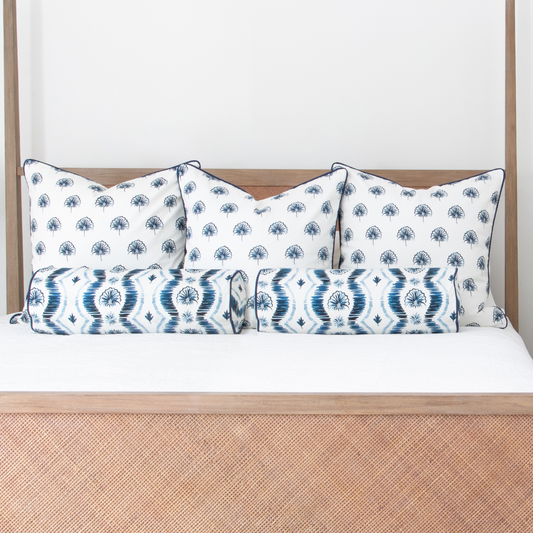 Blue ikat striped custom pillows and floral navy custom pillows styled on a light wood bed with white sheets in a room with white walls.