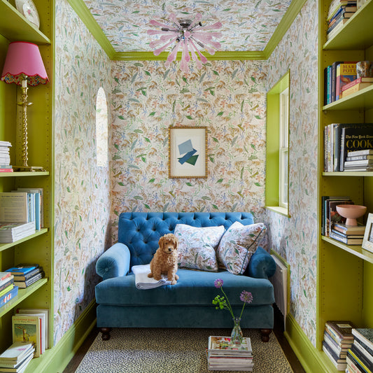 Library with pink chinoiserie wallpaper and chartreuse trim styled with a blue velvet couch and pink chinoiserie pillows next to a small dog