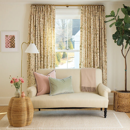 A cozy living room with floral curtains, a cream loveseat adorned with pastel pillows, a woven side table with fresh flowers, and a fiddle-leaf fig tree in a basket.