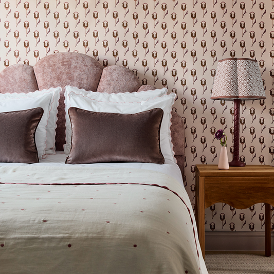 neutral background wallpaper with pink and maroon abstract floral pattern on it in a bedroom with a pink velvet headboard on the bed with white bedding and white pillows with brown velvet pillows and a brown wooden nightstand with a lamp on it