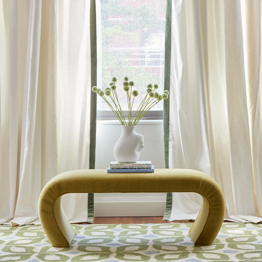 Natural white curtain with green velvet trim hanging on a window behind a chartreuse velvet bench with vase and green flowers