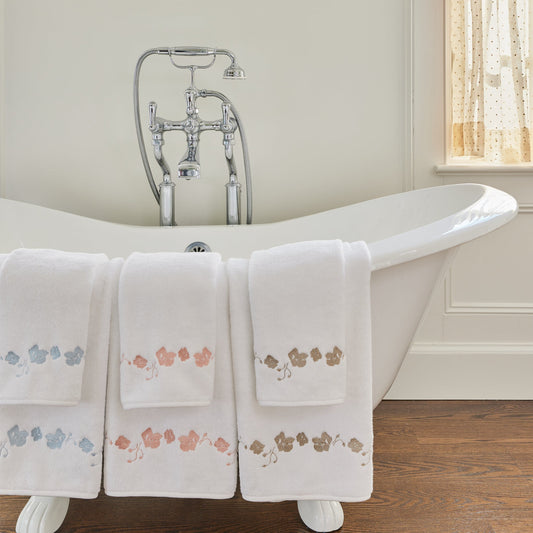 White towels with blue floral embroidered draped on a white bath tub in a bathroom with sky blue botanical wallpaper 