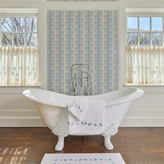 White tub with blue floral embroidered towels hanging on the side next to a matching bath mat in a bathroom with blue botanical wallpaper