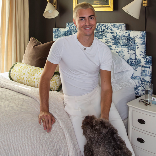 Blonde man wearing all white sitting on the corner of a white bed with one hand on a brown dog next to a dresser 