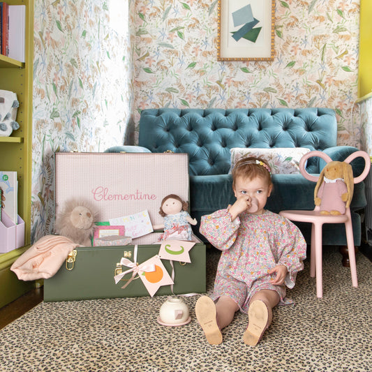 Little girl sitting on the floor in a pink dress next to a green trunk with pink fabric in a nook with pink chinoiserie tiger wallpaper and a blue velvet couch