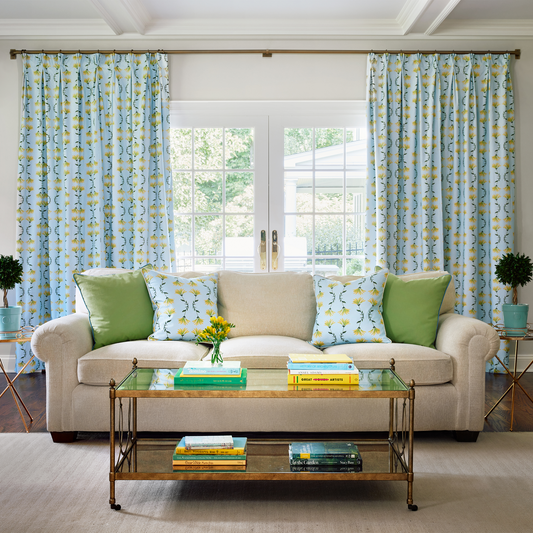 Living room with a clear coffee table in front of a beige couch styled with two green pillows and two blue botanical Ikat custom pillows in front of an illuminated window with blue botanical Ikat custom curtains 