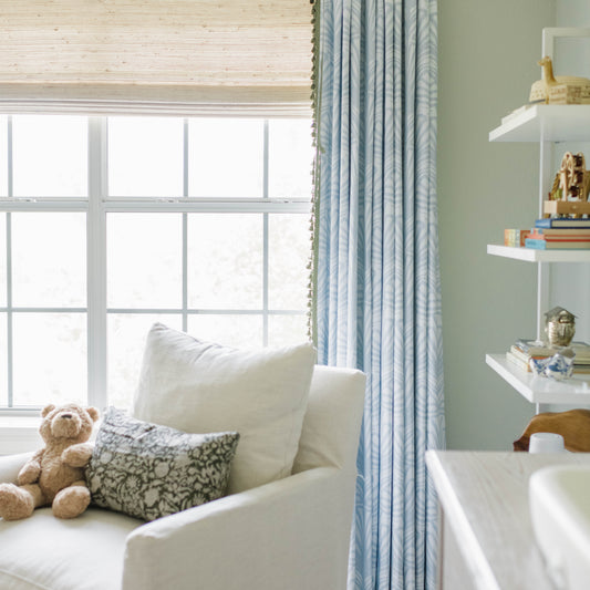 Blue palm curtain panel hanging on a window in a nursery behind a white chair with a teddy bear next to a bookshelf