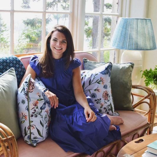 Brunette woman wearing a blue dress sitting on a pink couch in between two Daphne Powder custom pillows and two fern green velvet custom pillows 