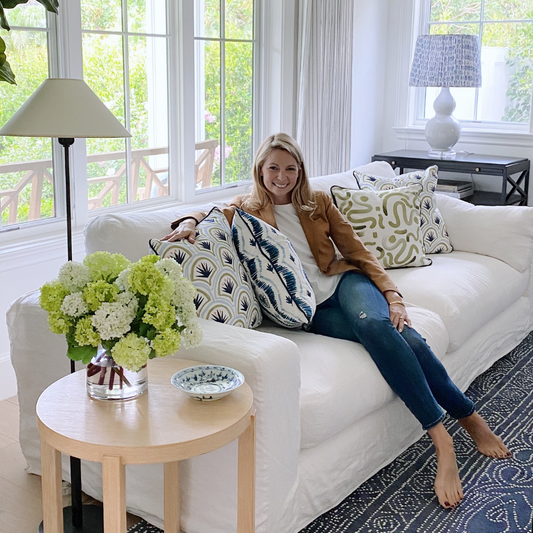 White couch with blonde woman sitting in between two art deco palm pattern custom pillows, a blue ikat stripe custom pillow, and a moss green custom pillow next to a circular wooden table with flowers in clear vase by tall lamp