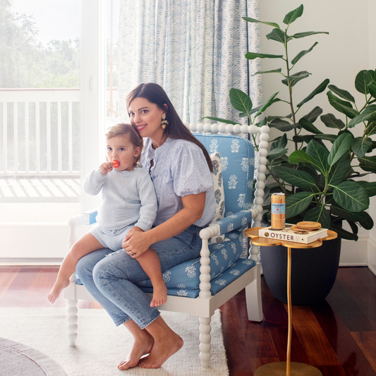 Brunette woman sitting on a blue and white armchair with child on lap next to plants in a big black clay vase in front of an illuminated window styled with Sky Blue Botanical Stripe Custom Curtains
