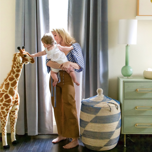 Blonde mother carrying baby touching a big stuffed giraffe in front of an illuminated window styled with Sky Blue Velvet Custom Curtains