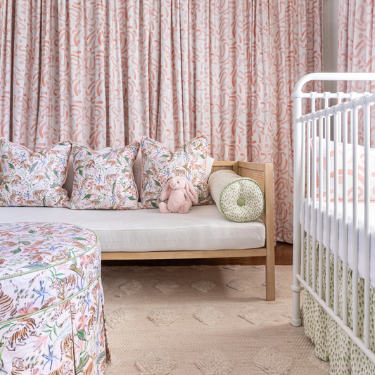 Nursery room corner styled with Pink Chinoiserie Tiger Custom Pillows on wooden bench with white mattress matching with a Pink Chinoiserie Tiger Custom Ottoman with Pink Graphic Custom Curtains next to a white crib with a pink graphic custom lumbar pillow