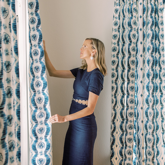 Blonde woman wearing a navy blue dress in front of an illuminated window holding a Blue Ikat Stripe Custom Curtain next to another Blue Ikat Stripe Custom Curtain on a window behind 
