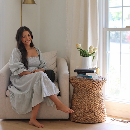 Brunette woman wearing a long blue dress sitting in white sofa chair next to an illuminated window with natural white custom curtains and a small table next to it with white tulips in white vase by stacked books on top