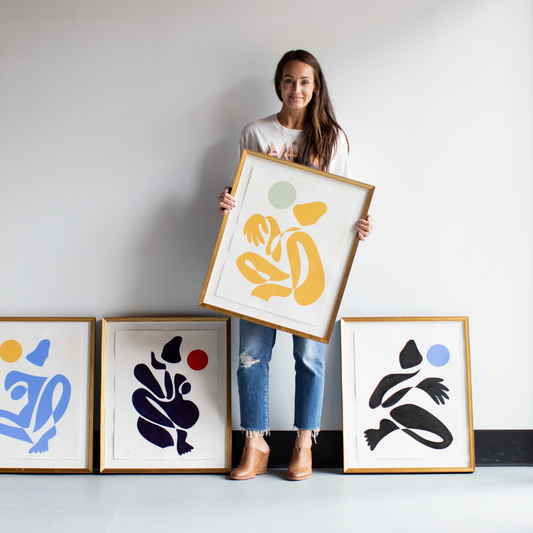 Brunette woman standing in front of a white wall holding her yellow modern artwork next to black and blue modern artwork