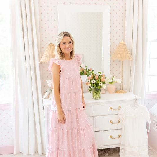 Blonde woman standing on front of white dresser in a pink and white nursery with natural white curtains hanging on two windows