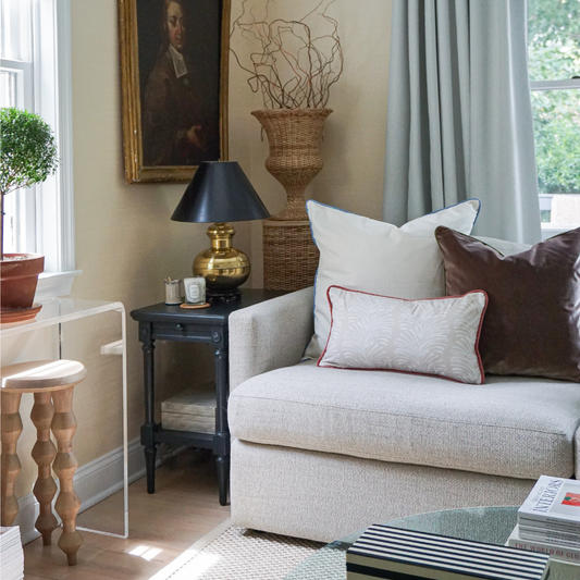 Living room corner styled with a Natural White Linen Custom Pillow, a Brown Velvet Custom Pillow, and a Beige Botanical Stripe Custom Lumbar on couch by black table with gold and black lamp on it