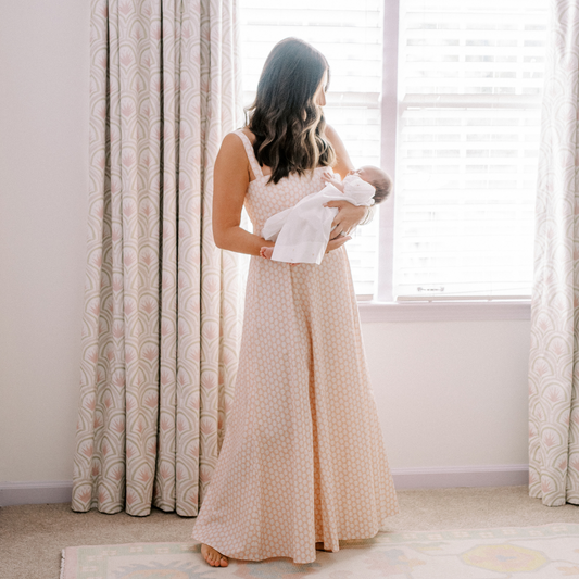 Brunette woman in pink dress holding a newborn baby in front of a window with pink and green art deco curtain panels