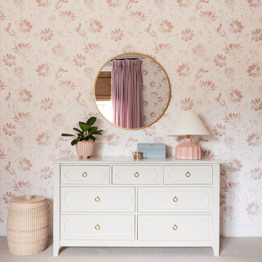 Bedroom with pink floral wallpaper styled with a white dresser, rattan basket, and a pink lamp below a gold mirror