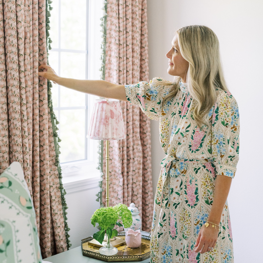 Close-Up of Bedroom Corner with Pink Floral Curtains with Sage Tassel being touched by blonde woman in a flower printed dress in front of illuminated window