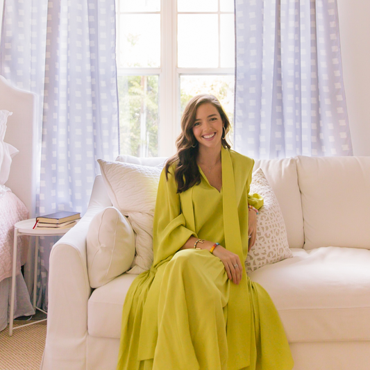 woman sitting on a white chair in front of a white desk and a window with sky blue patterned curtains hanging
