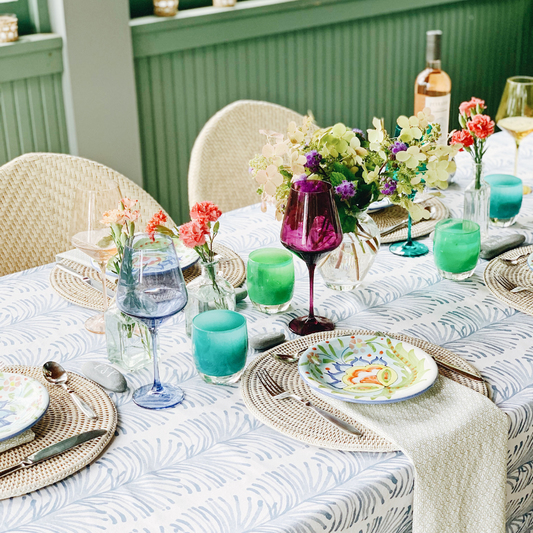 Table with rattan chairs styled with a sky blue botanical tablecloth and green and blue geometric napkins and rattan placemats and colorful floral plates