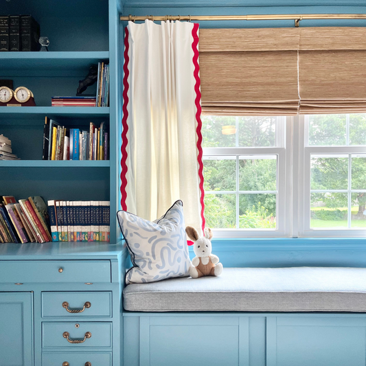 A blue painted room with a blue desk and bookshelf attached to a bench seat with blue fabric below a window with a white curtain panel with red rick rack trim paired with a rattan woven shade