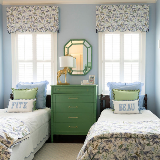 A blue painted kids room with two windows styled with blue and green tiger chinoiserie scalloped roman shades above two twin beds with white bedding and blue and green tiger duvet covers with a green dresser in between