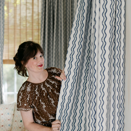 Brunette woman in a brown dress standing behind a blue and white squiggle embroidered curtain panel holding the sides with her hands