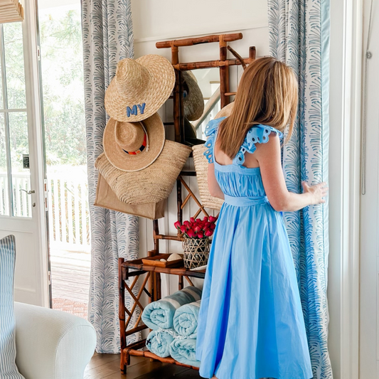 Woman wearing a long blue dress holding Sky Blue Botanical Stripe custom curtains next to a wooden hanger stand with beach hats, totes, and towels 