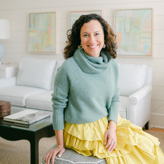 Brunette woman in a blue sweater and yellow skirt sitting on blue stool in a living room in front of a white couch and colorful artwork