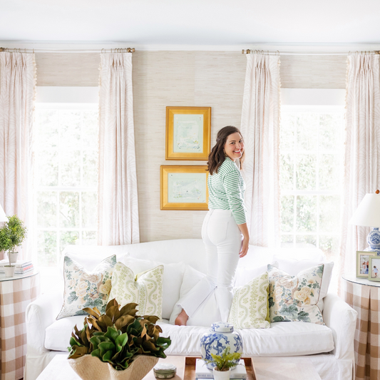 Living room with a brunette woman standing on top of a white couch with two illuminated windows styled with Beige Palm custom curtains on them