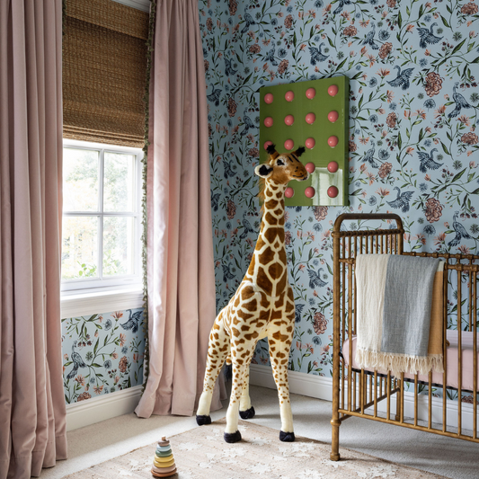 Corner of a nursery with blue chinoiserie wallpaper and one window styled with a rattan roman shade and pink velvet custom curtains next to a brass crib and a large stuffed animal girafe