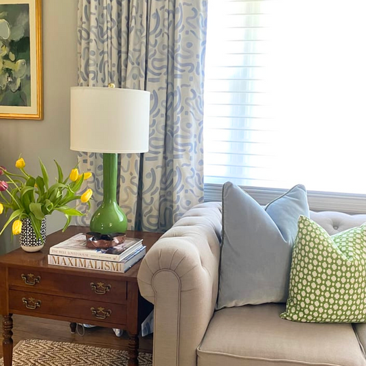 Living room corner with a beige couch styled with Sky Blue Velvet Custom Pillow and green and white pillow next to a wooden table with a green lamp, flowers in vase, and stacked books on top by Abstract Sky Blue Custom Curtains 