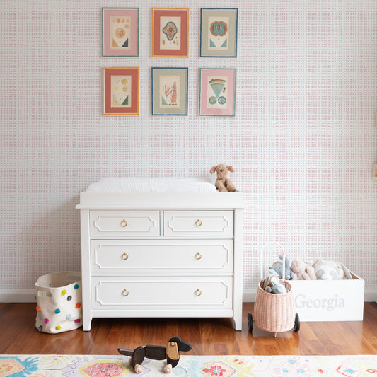 Nursery close up of a white changing table below colorful artwork on a wall with rose gingham wallpaper 