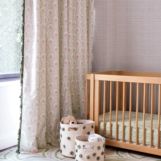 Corner of a nursery with rose pink gingham wallpaper and pink art deco palm curtains hanging in front of a window next to a wooden crib with green crib sheets and white toy baskets with green pom poms