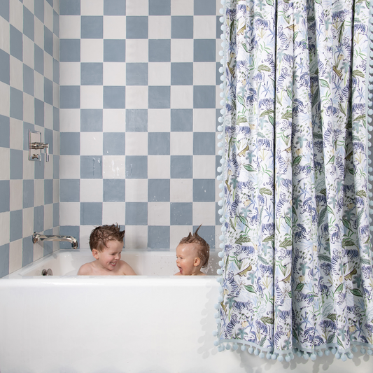 Green tiger shower curtain hanging in front of a white tub in bathroom with blue and white tiles and two young boys playing