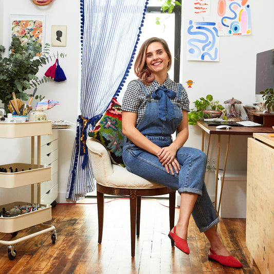 Brunette in overalls sitting on a chair at her desk in her art studio with an art supplies cart at her side and whimsical artwork hanging on the walls