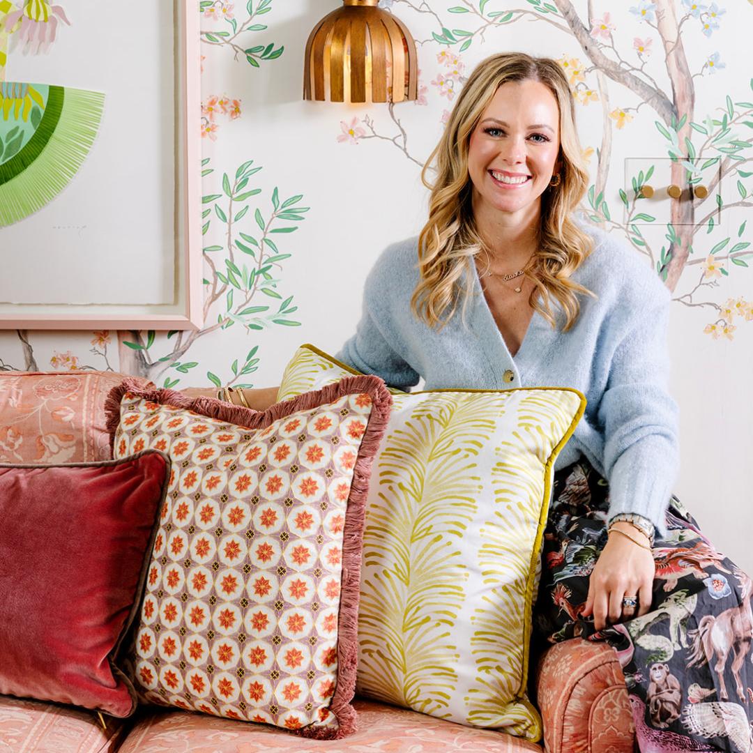 A woman sits on a pink patterned couch styled with a chartreuse and white botanical pillow with yellow piping, a fringed floral print pillow in soft red and cream, and a rich reddish-pink velvet pillow, in a floral-themed room. 