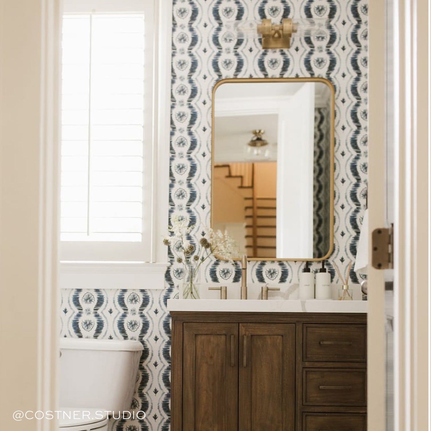 Bathroom with Blue Ikat Striped Pattern Wallpaper with gold framed mirror on top of white sink on wooden doors and three drawers by white toilet under illuminated white window