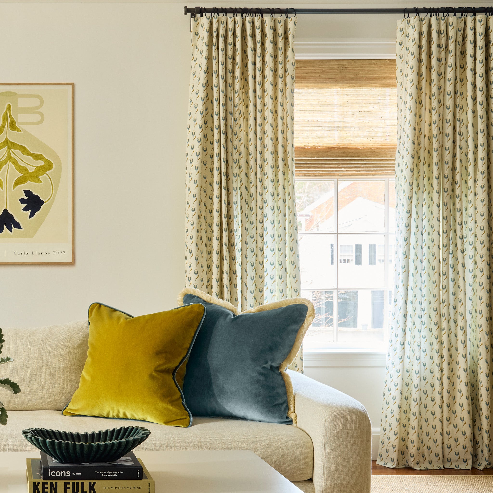 Abstract floral blue and green printed curtains on a metal rod in front of an illuminated window with a white couch with golden chartreuse velvet and peacock blue velvet pillows on it and a coffee table stacked with books