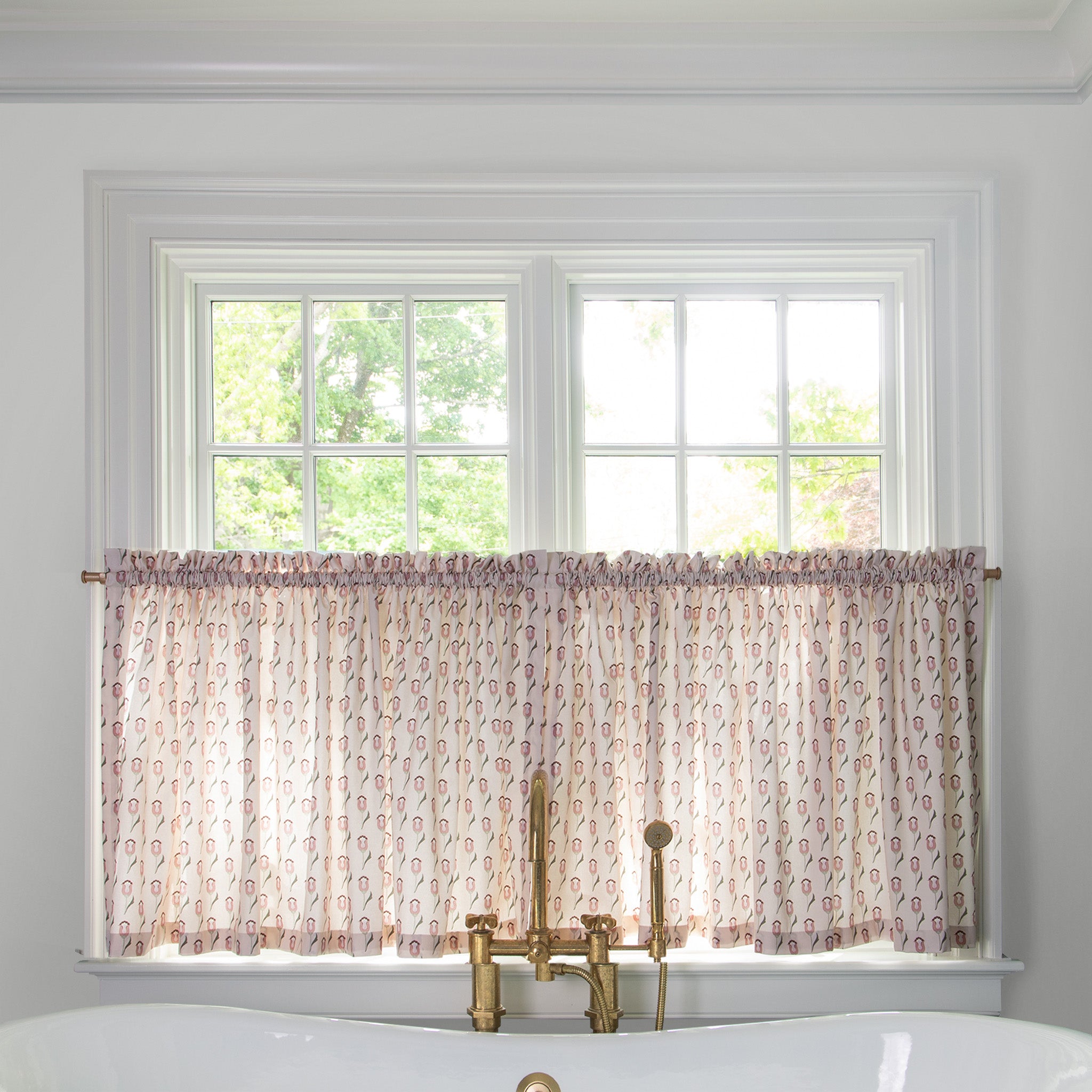 light pink fabric with pink abstract floral pattern cafe curtain hung in front of an illuminated window in a bathroom with a white bathtub in front of the window with a gold faucet 