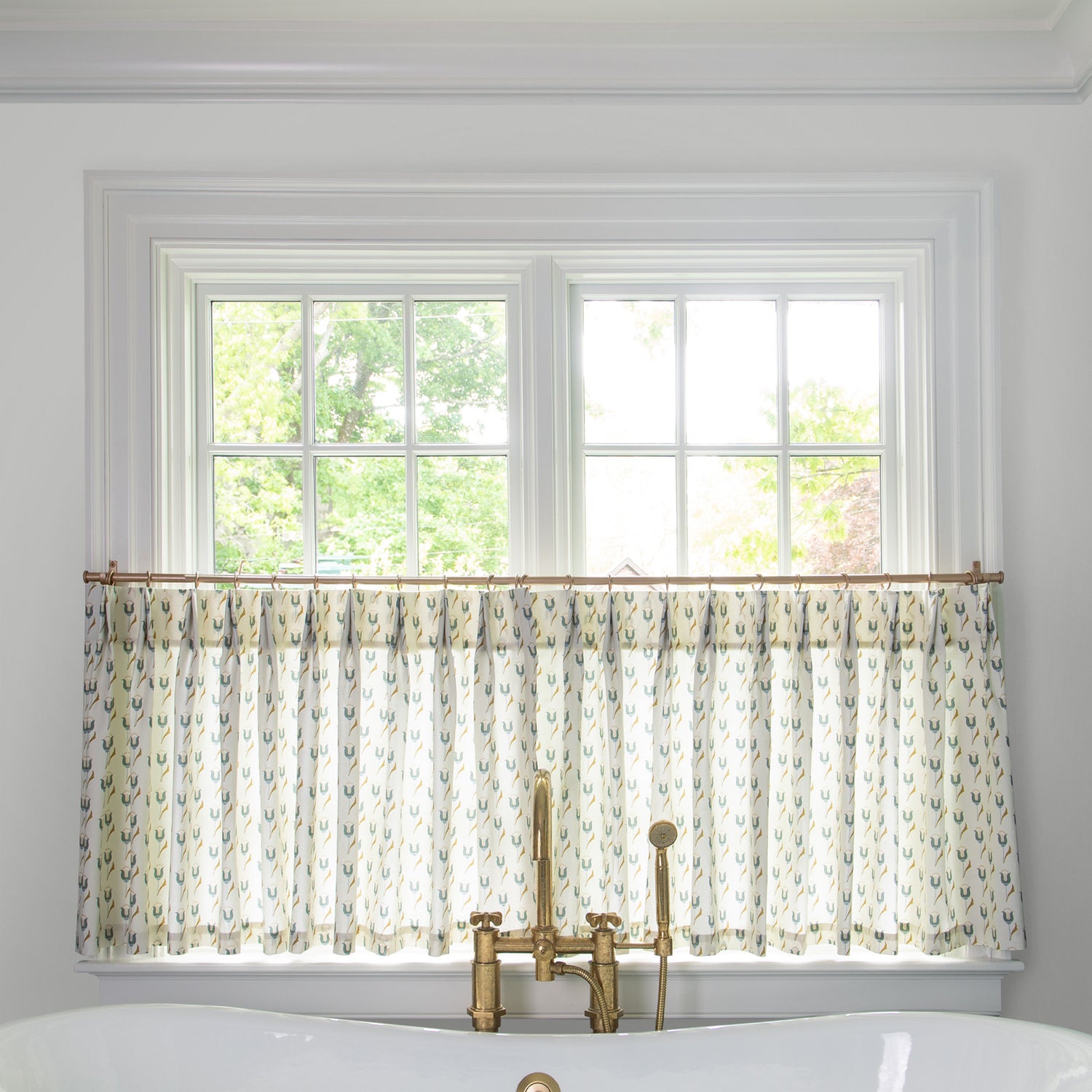 neutral fabric with green abstract floral pattern cafe curtain hung in front of an illuminated window in a bathroom with a white bathtub in front of the window with a gold faucet 